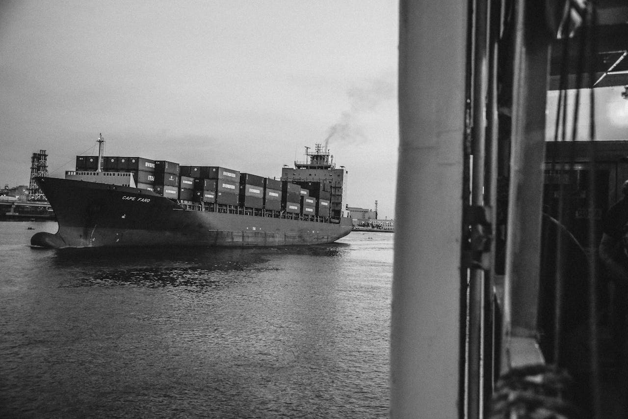 Black and white image of a cargo ship with containers, moving through a harbor during the day.